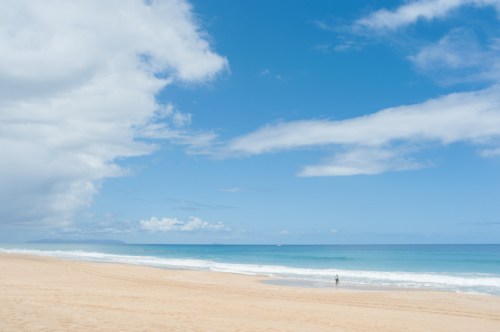 A serene beach scene with soft sand, gentle waves, and a clear blue sky dotted with clouds. A lone figure stands by the water.