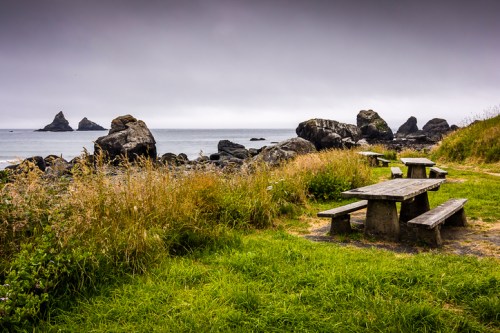 A coastal scene featuring a picnic table near rocky formations and a cloudy sky, surrounded by tall grass.