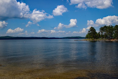 A serene lake scene with clear water, lush trees, and fluffy clouds under a bright blue sky.