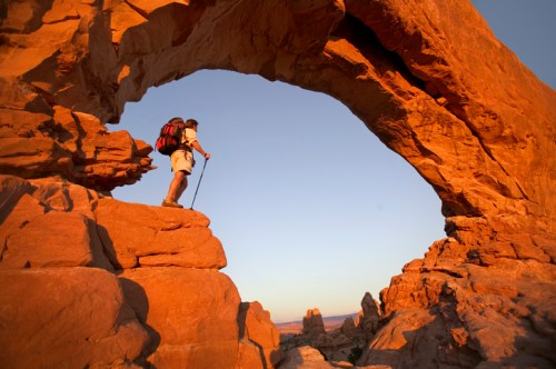 A hiker stands at the edge of a rock arch, overlooking a desert landscape at sunset.