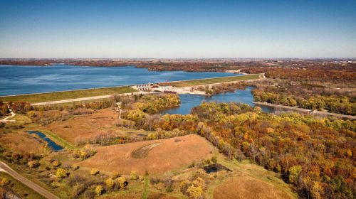 Aerial view of a lake and dam surrounded by autumn foliage and fields under a clear blue sky.