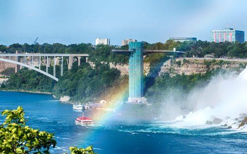 A view of Niagara Falls with a rainbow over the water, featuring a boat and a tall observation tower in the background.
