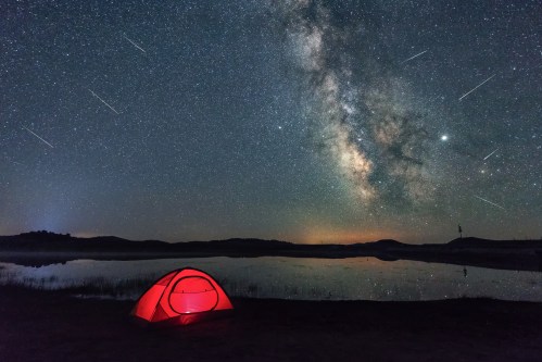 A red tent by a calm lake under a starry sky filled with the Milky Way and shooting stars.