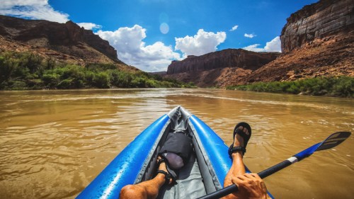 A person paddles a kayak on a river surrounded by rocky cliffs and blue skies with fluffy clouds.