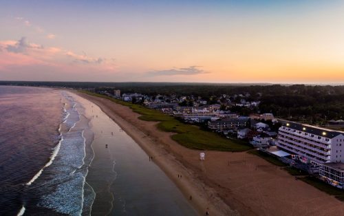 Aerial view of a beach at sunset, with waves, sandy shore, and a small town in the background.
