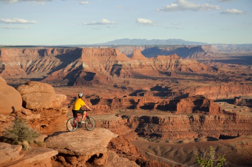 A cyclist in a yellow shirt overlooks a vast canyon landscape with red rock formations and distant mountains.