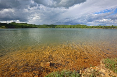 A serene lake with clear water, rocky shore, and lush green hills under a cloudy sky.