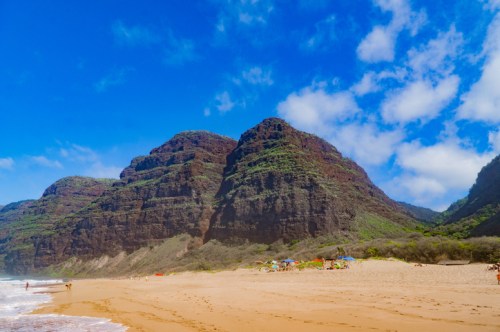 A scenic beach with golden sand, lush green mountains, and a bright blue sky with scattered clouds.