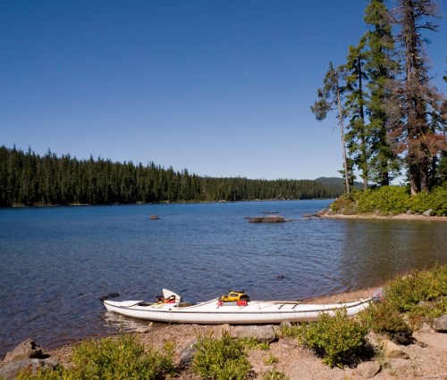 A calm lake surrounded by trees, with a white kayak resting on the shore under a clear blue sky.
