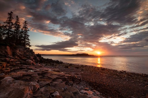 Sunset over a rocky shoreline with trees silhouetted against a colorful sky and calm water.
