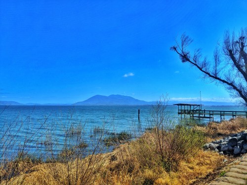 A serene lakeside view with a wooden pier, mountains in the distance, and clear blue skies.
