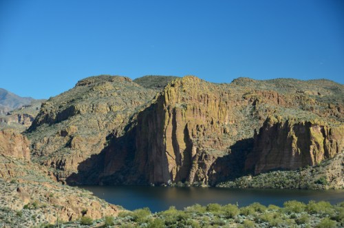 A scenic view of rugged cliffs and a calm lake under a clear blue sky, surrounded by desert vegetation.