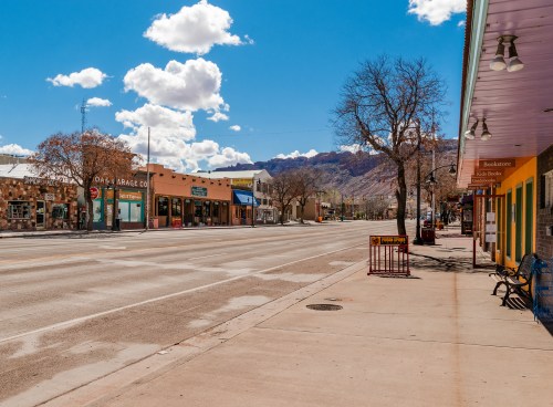 A quiet street scene with shops and mountains in the background, under a bright blue sky with fluffy clouds.