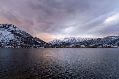 A serene lake surrounded by snow-capped mountains under a cloudy sky at dusk.