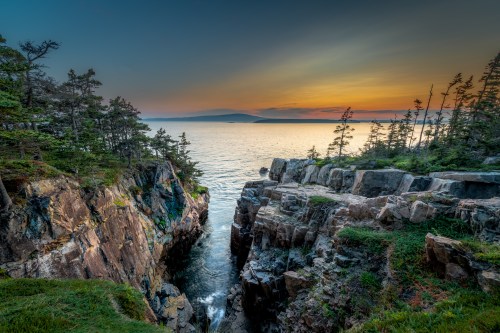 A serene coastal scene at sunset, featuring rocky cliffs, lush trees, and calm waters reflecting warm hues.
