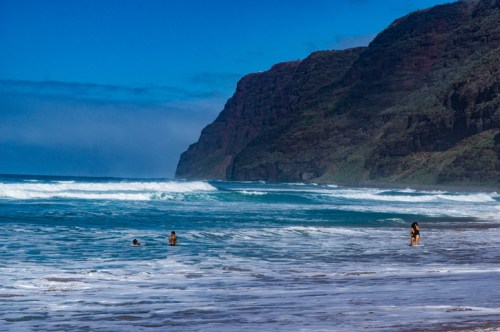 Two people wade in the ocean while another stands on the shore, with cliffs and blue skies in the background.