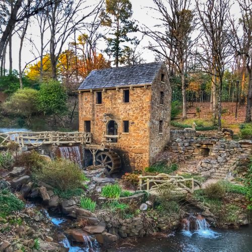 A historic stone mill with a water wheel, surrounded by trees and a flowing stream, in a serene outdoor setting.