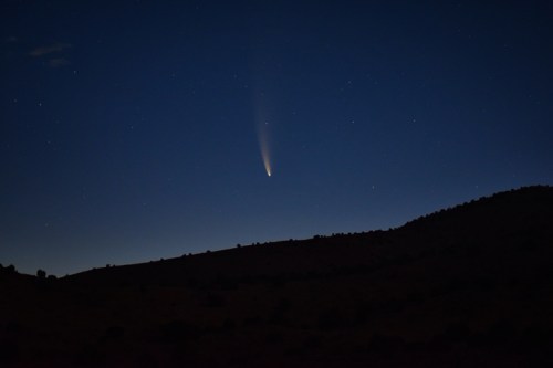 A comet streaks across a dark night sky filled with stars, silhouetting a distant mountain range.