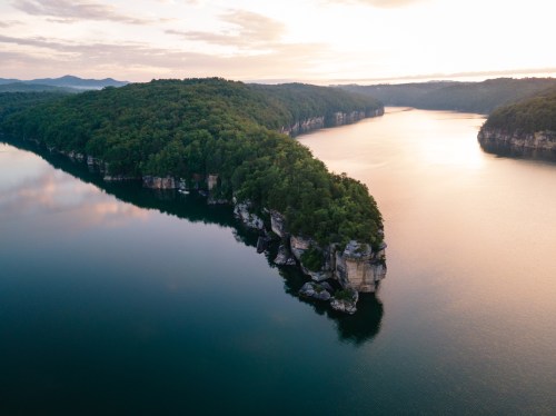 Aerial view of a serene lake surrounded by lush green hills and rocky cliffs at sunset.