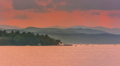 A serene lake at sunset, with mountains in the background and boats scattered on the water under a colorful sky.