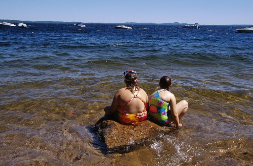Two children sit on a rock by the water, wearing colorful swimsuits, enjoying a sunny day at the lake.