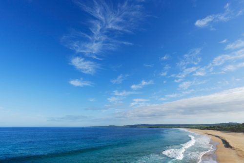A serene beach scene with gentle waves, a clear blue sky, and wispy clouds above the coastline.