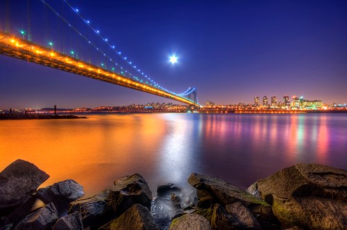 A vibrant night scene of a bridge illuminated by lights, reflecting on calm waters with a city skyline in the background.
