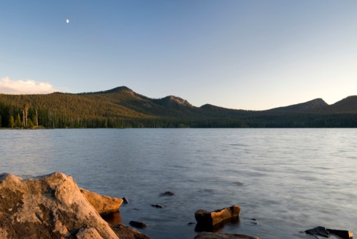 A serene lake surrounded by mountains and trees, with a clear sky and a crescent moon visible.