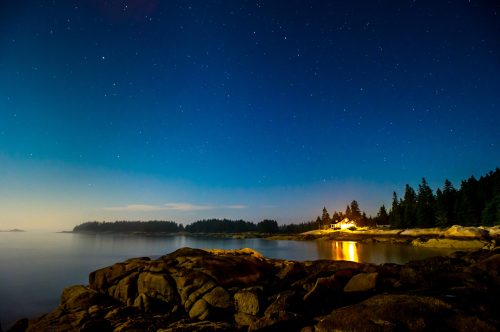 A serene night scene with stars above, reflecting on calm water near rocky shores and a distant lit cabin among trees.