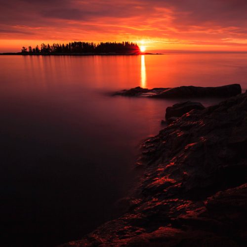 A serene sunset over calm waters, with a silhouette of trees on an island and rocky shoreline in the foreground.