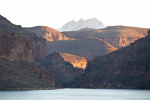 Sunlit mountains rise above a serene lake, surrounded by rugged cliffs and lush greenery in a tranquil landscape.