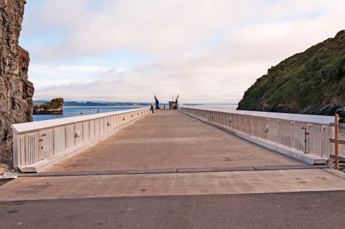 A wide, empty pier extends into calm waters, flanked by rocky cliffs and greenery under a cloudy sky.