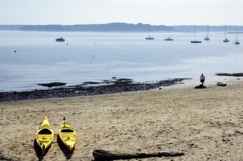 Two yellow kayaks on a sandy beach with boats in the calm water and a person walking along the shore.