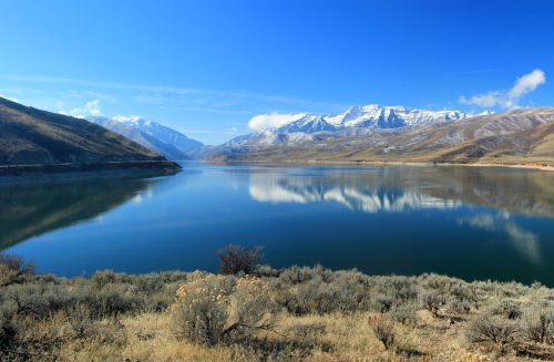 A serene lake reflects snow-capped mountains under a clear blue sky, surrounded by dry shrubs and hills.