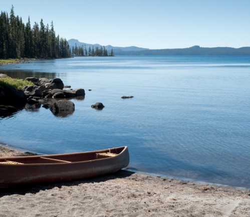 A calm lake scene with a canoe on the sandy shore, surrounded by trees and distant mountains under a clear blue sky.