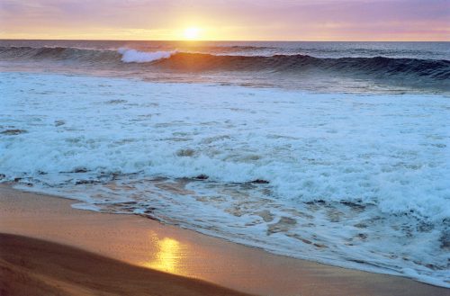 Sunset over the ocean, with waves gently crashing on a sandy beach, reflecting warm colors in the water.