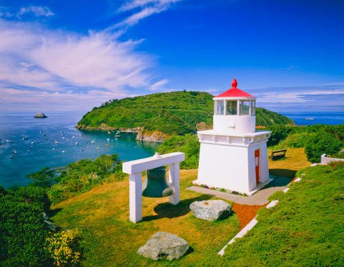 A scenic view of a lighthouse on a hill overlooking a calm bay with boats and lush greenery under a blue sky.