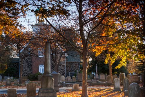 A serene cemetery scene with autumn leaves, gravestones, and a historic building in the background.