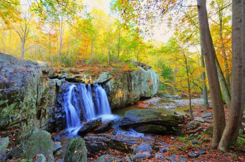 A serene waterfall cascades over rocks, surrounded by vibrant autumn foliage and tranquil forest scenery.