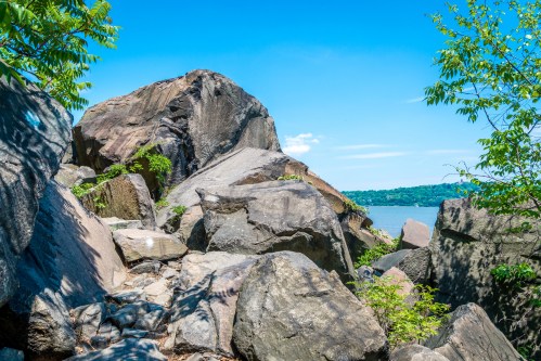 Rocky landscape with large boulders and greenery, overlooking a calm body of water under a clear blue sky.