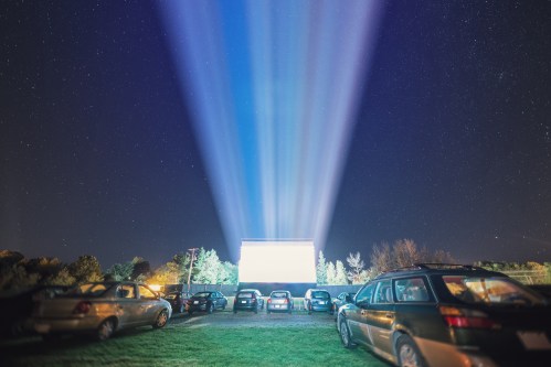 A night scene at a drive-in theater with cars parked and beams of light shining from a large screen under a starry sky.