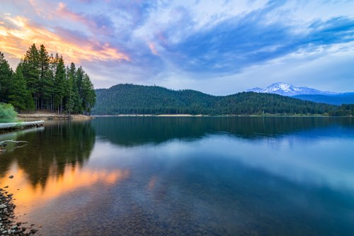 A serene lake at sunset, surrounded by trees and mountains, with colorful reflections on the water's surface.
