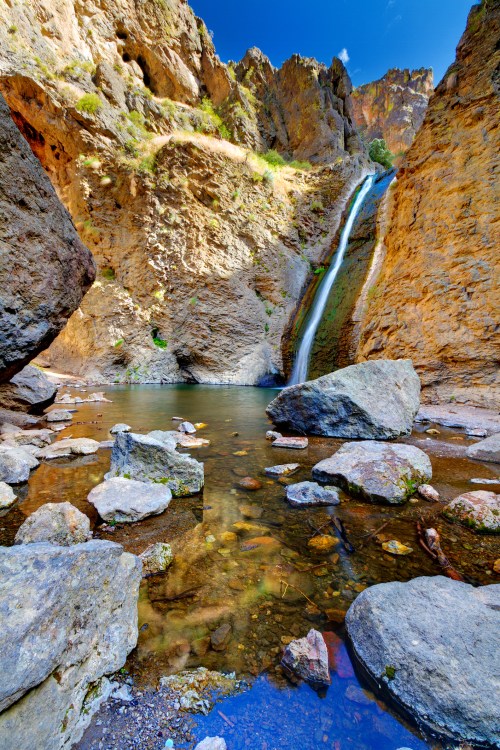 A serene waterfall cascades into a clear pool, surrounded by rocky cliffs and vibrant blue sky.