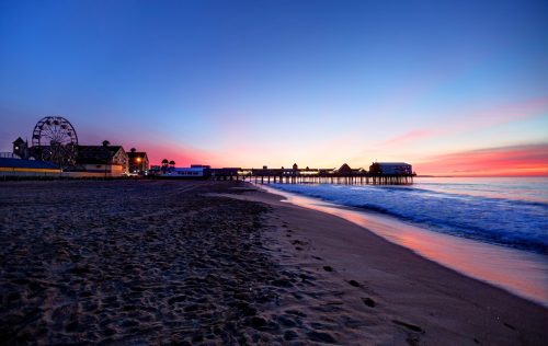A serene beach at sunset, featuring a pier and a ferris wheel, with colorful skies reflecting on the water.