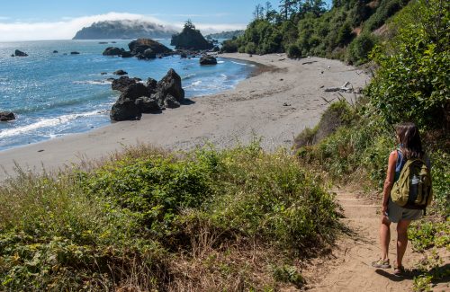 A hiker descends a path towards a sandy beach with rocky formations and a misty coastline in the background.