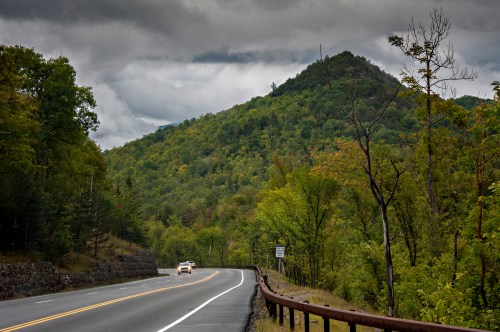 A winding road leads through lush green hills under a cloudy sky, with a car driving in the distance.