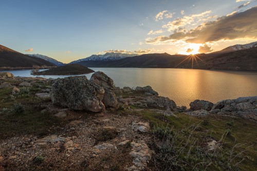 A serene lake at sunset, surrounded by mountains and rocky terrain, with sunlight reflecting on the water's surface.