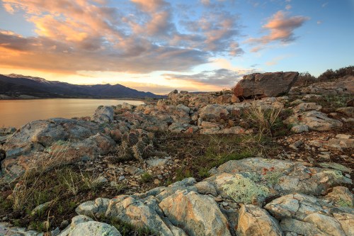 Scenic view of rocky shoreline at sunset, with mountains and colorful clouds reflected in the water.