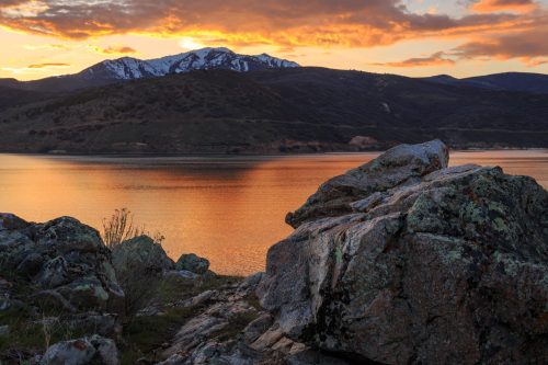 A serene lake at sunset, with mountains in the background and rocks in the foreground reflecting warm colors.