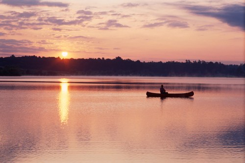 A person in a canoe paddles on a calm lake at sunset, with soft clouds reflecting on the water's surface.